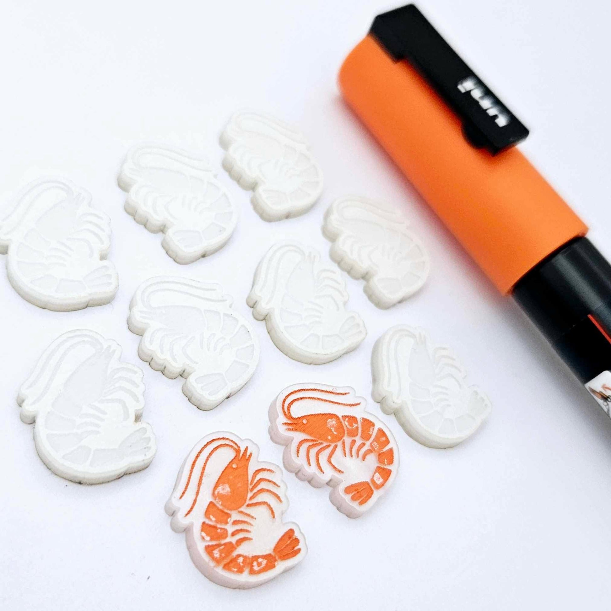 One paint-filled pair of White Marble acrylic prawn stud earring blanks with multiple unpainted pieces still covered with protective paper, pictured alongside an Orange Posca Paint Pen