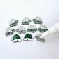 One paint-filled pair of green glitter acrylic Santa hat toppers alongside multiple unpainted pieces still covered with protective paper, pictured with an uncapped White Posca Paint Pen ready for paint filling