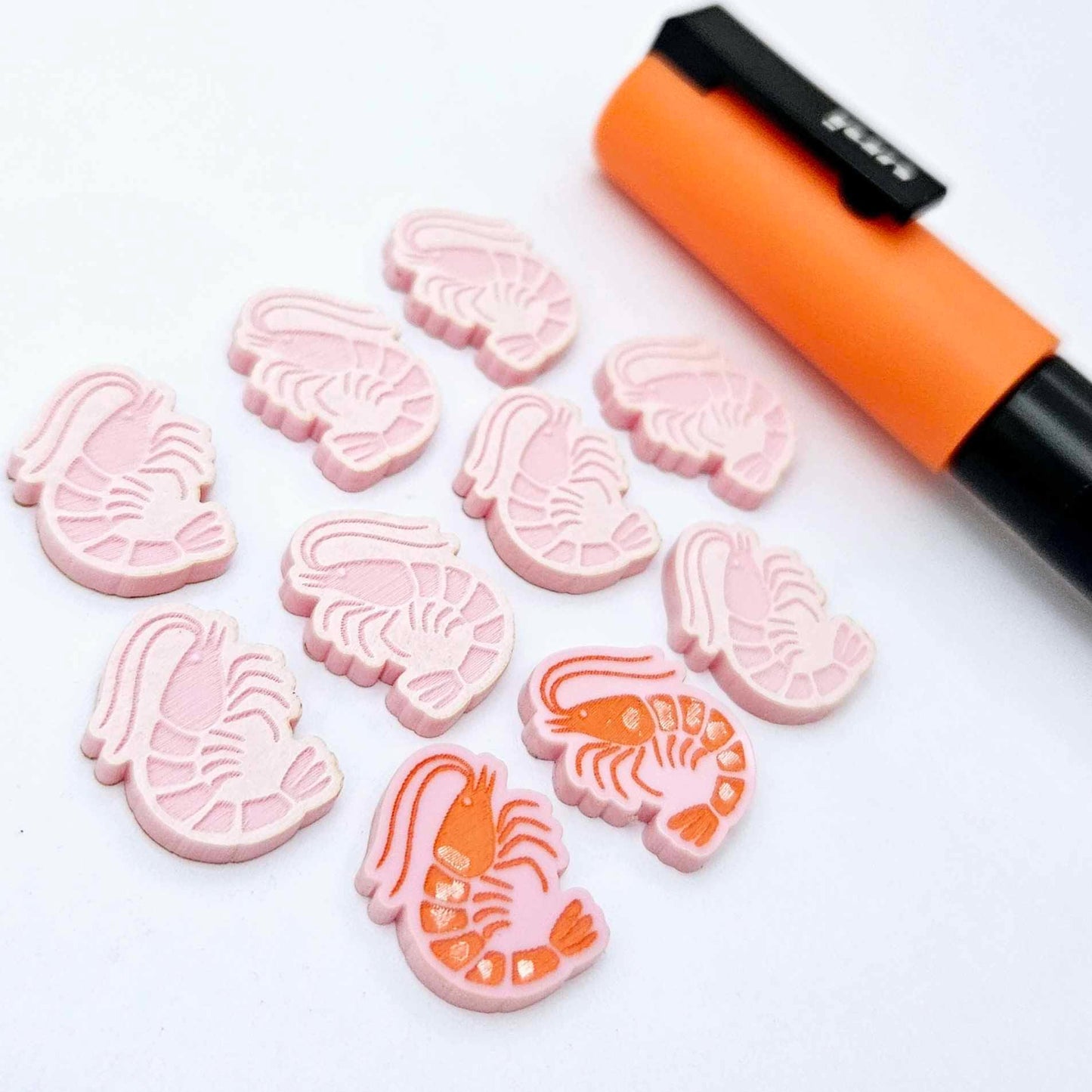 One paint-filled pair of pastel pink acrylic prawn stud earring blanks with multiple unpainted pieces still covered with protective paper, pictured alongside an Orange Posca Paint Pen