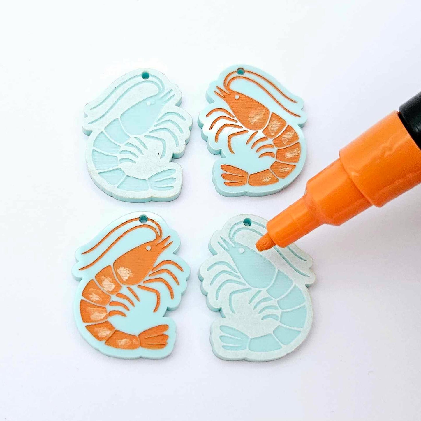 One pair of pastel aqua prawn dangle blanks with protective paper attached, alongside one paint-filled pair with a posca paint pen uncapped ready to paint