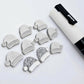 One paint-filled pair of silver glitter acrylic Santa hat stud blanks alongside multiple unpainted pieces still covered with protective paper, pictured with a White Posca Paint Pen