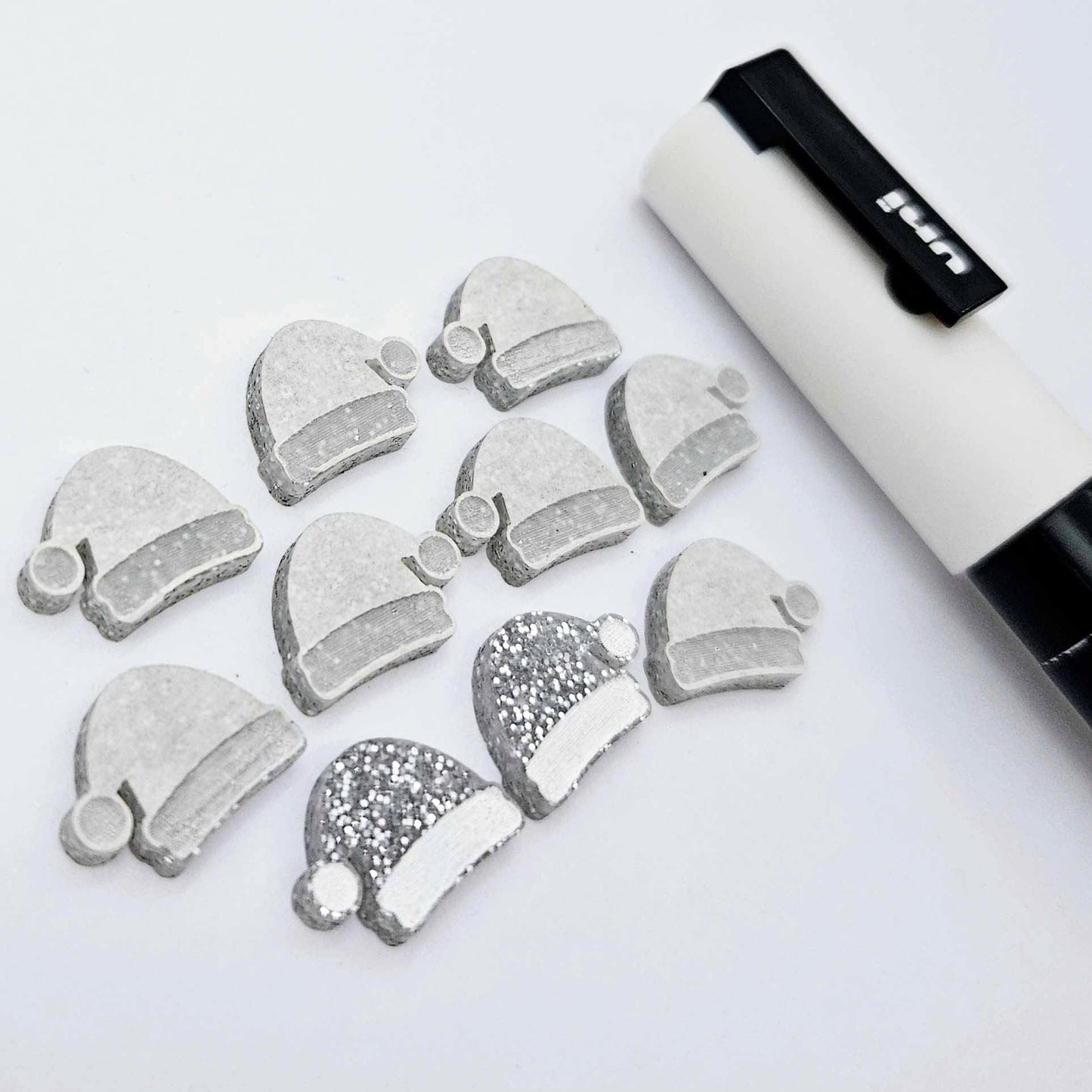 One paint-filled pair of silver glitter acrylic Santa hat stud blanks alongside multiple unpainted pieces still covered with protective paper, pictured with a White Posca Paint Pen