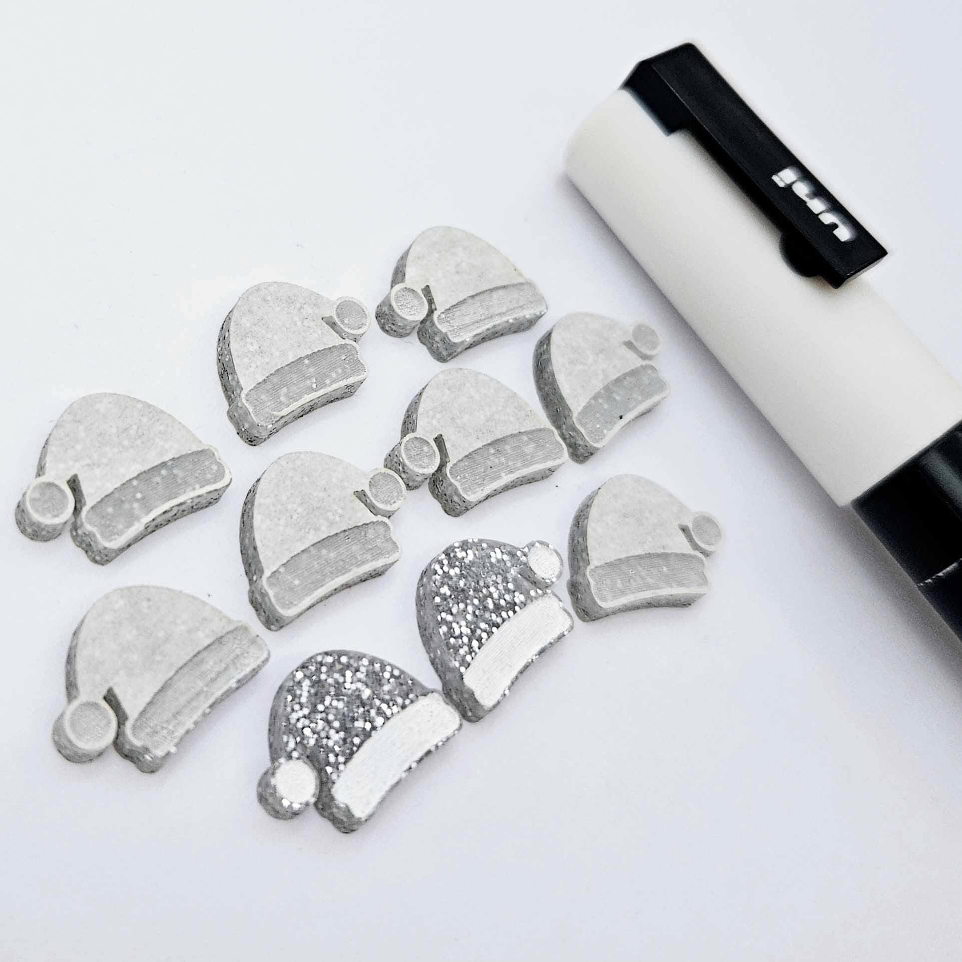 One paint-filled pair of silver glitter acrylic Santa hat stud blanks alongside multiple unpainted pieces still covered with protective paper, pictured with a White Posca Paint Pen