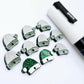 One paint-filled pair of green glitter acrylic Santa hat earring toppers alongside multiple unpainted pieces still covered with protective paper, pictured with a White Posca Paint Pen