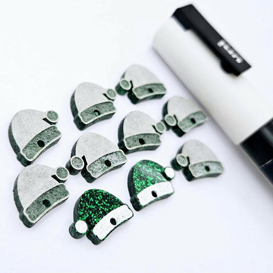 One paint-filled pair of green glitter acrylic Santa hat earring toppers alongside multiple unpainted pieces still covered with protective paper, pictured with a White Posca Paint Pen