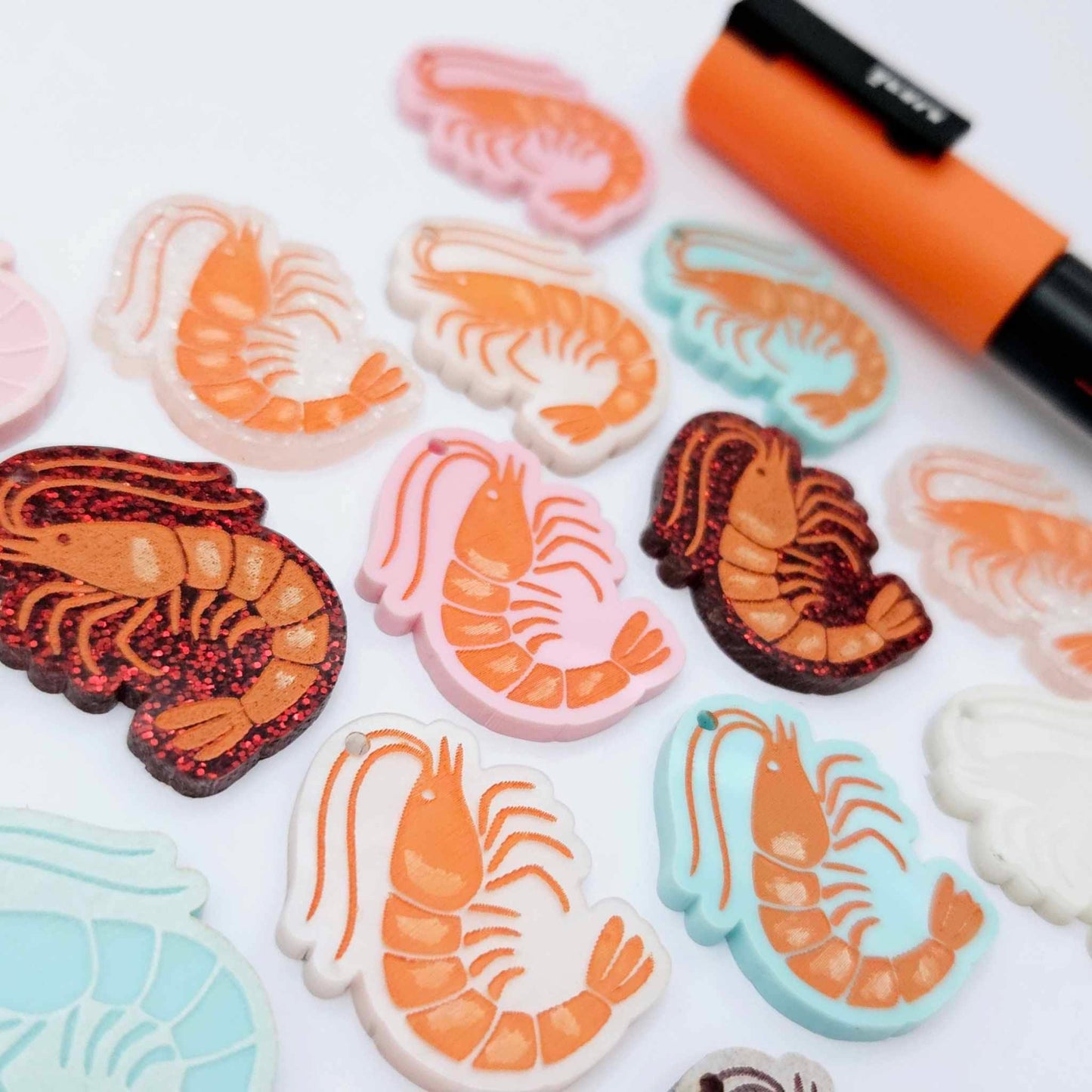 A mixture of Pastel Pink, Pastel Aqua, Red Glitter, Fairy Dust Glitter and White Marble acrylic prawn dangle earring blanks laying flat with an orange posca paint pen