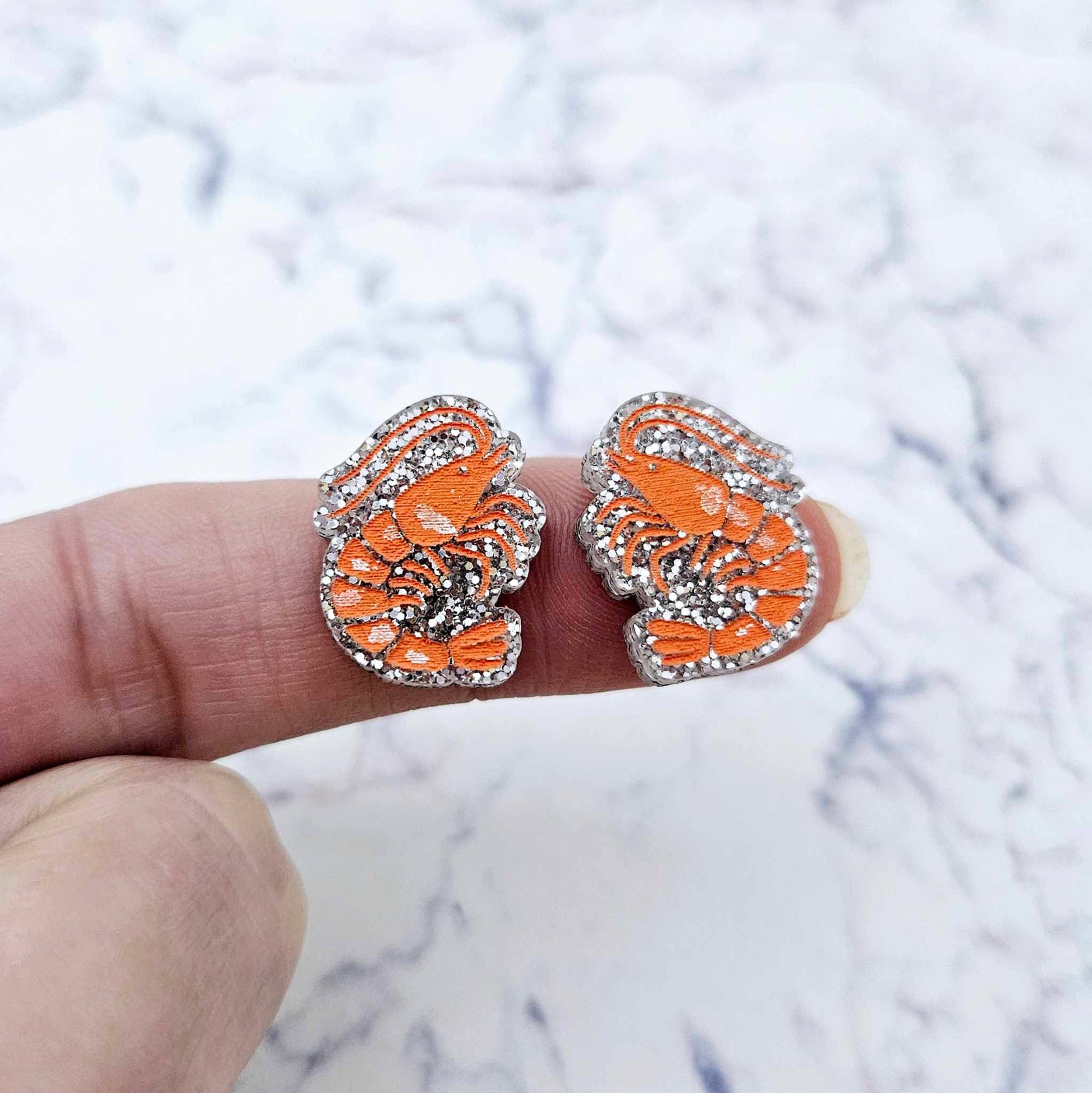 One pair of paint-filled silver glitter acrylic prawn stud earring blanks shown on a finger for size reference