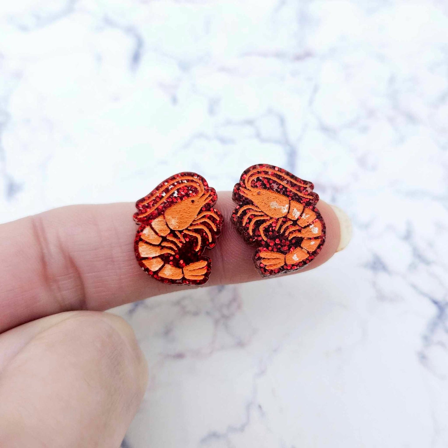 One pair of paint-filled red glitter acrylic prawn stud earring blanks shown on a finger for size reference