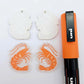 One pair of fairy dust glitter acrylic engraved prawn dangle earring blanks with protective paper on alongside a paint filled pair, laying flat with an orange posca paint pen