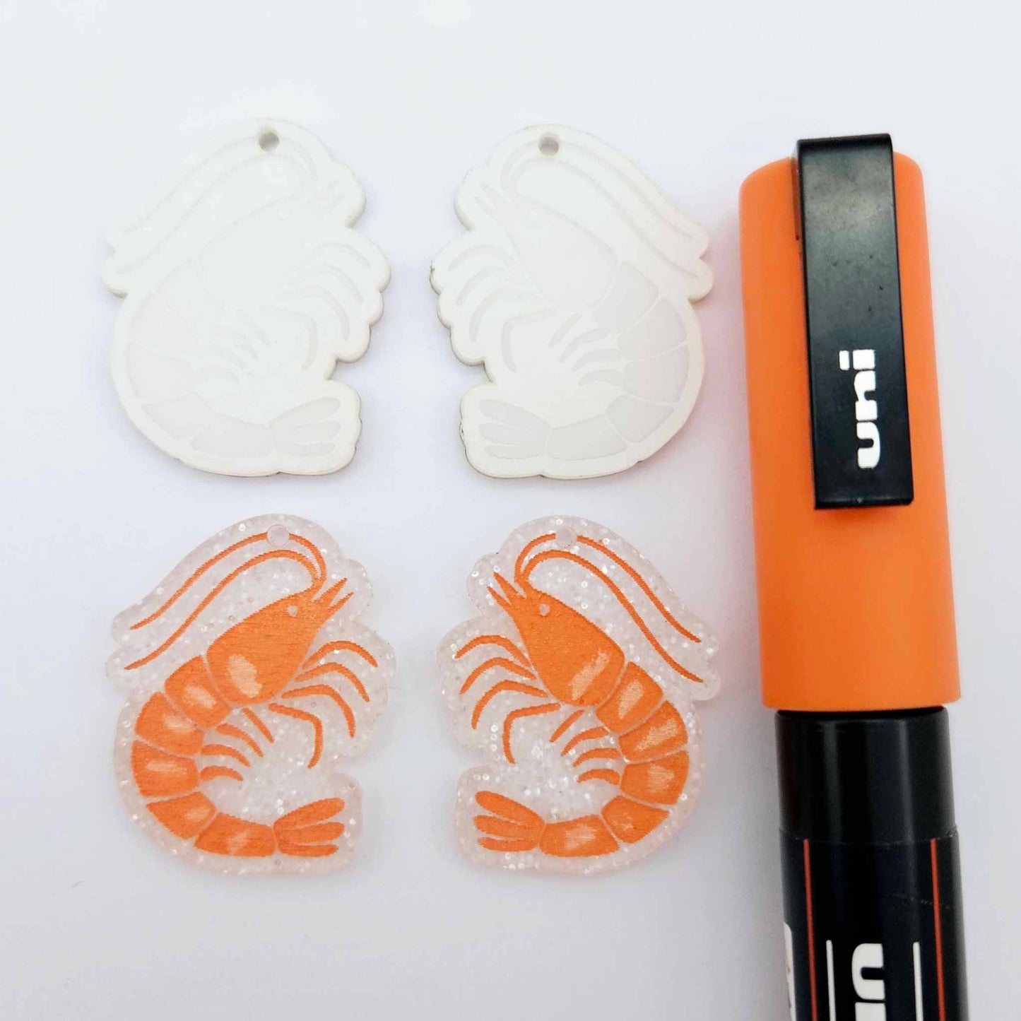 One pair of fairy dust glitter acrylic engraved prawn dangle earring blanks with protective paper on alongside a paint filled pair, laying flat with an orange posca paint pen