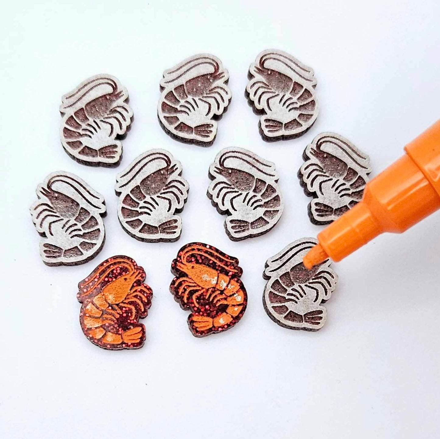 One paint-filled pair of red glitter acrylic prawn stud earring blanks with multiple unpainted pieces still covered with protective paper, pictured with an uncapped Orange Posca Paint Pen ready for paint filling