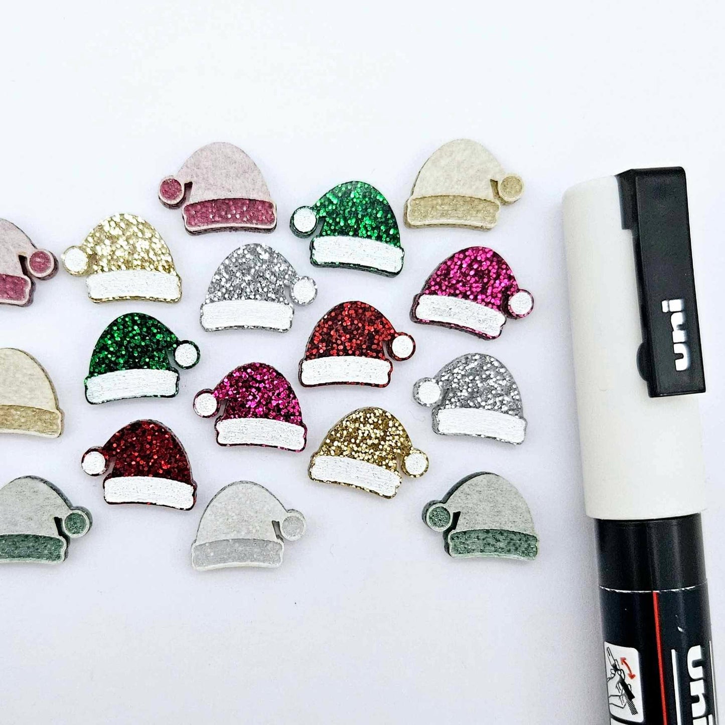A selection of paint-filled glitter acrylic Santa hat stud blanks in red, green, gold, silver and hot pink, with multiple unpainted pieces still covered with protective paper, pictured alongside a White Posca Paint Pen