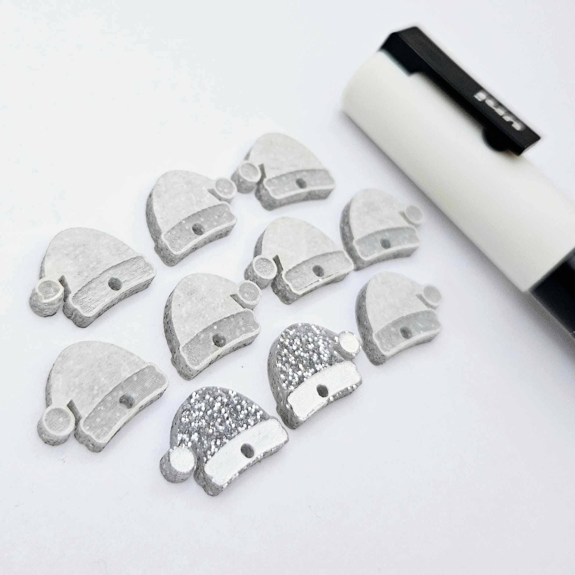 One paint-filled pair of silver glitter acrylic Santa hat toppers alongside multiple unpainted pieces still covered with protective paper, pictured with a White Posca Paint Pen