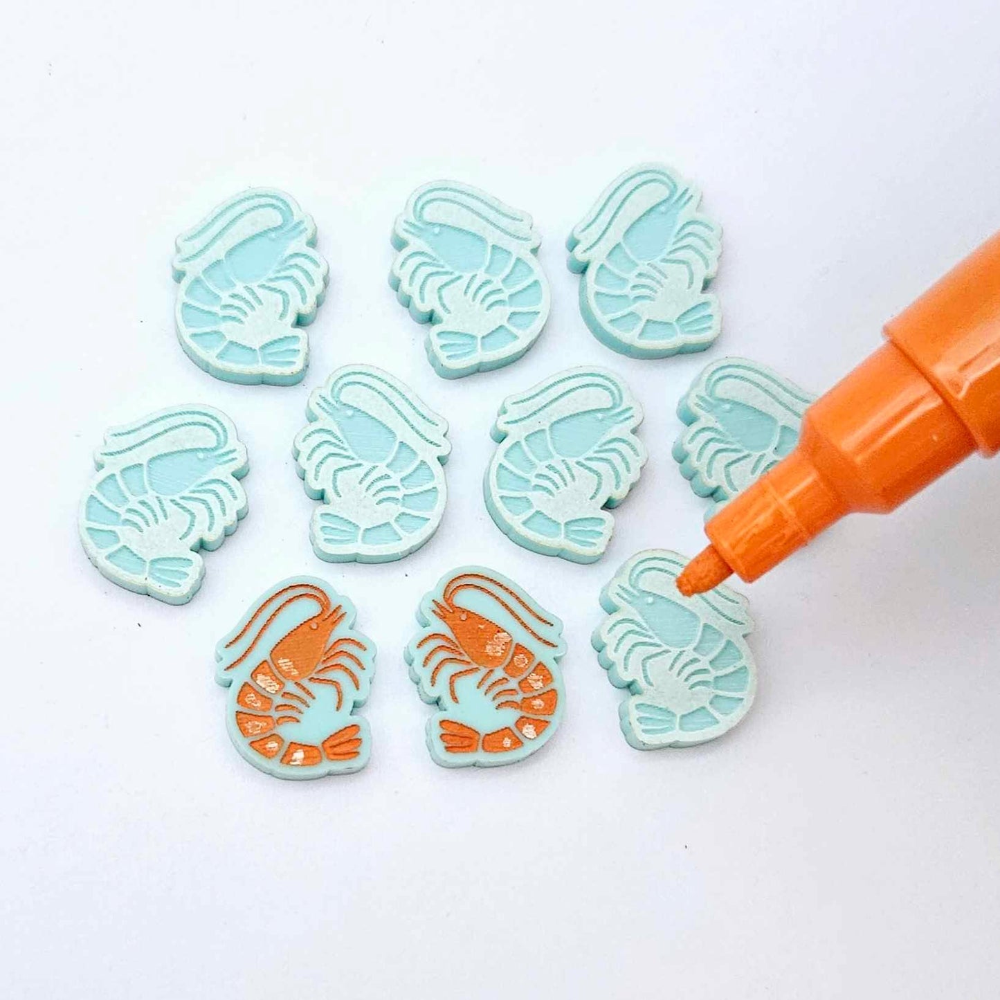 One paint-filled pair of pastel aqua acrylic prawn stud earring blanks with multiple unpainted pieces still covered with protective paper, pictured with an uncapped Orange Posca Paint Pen ready for paint filling