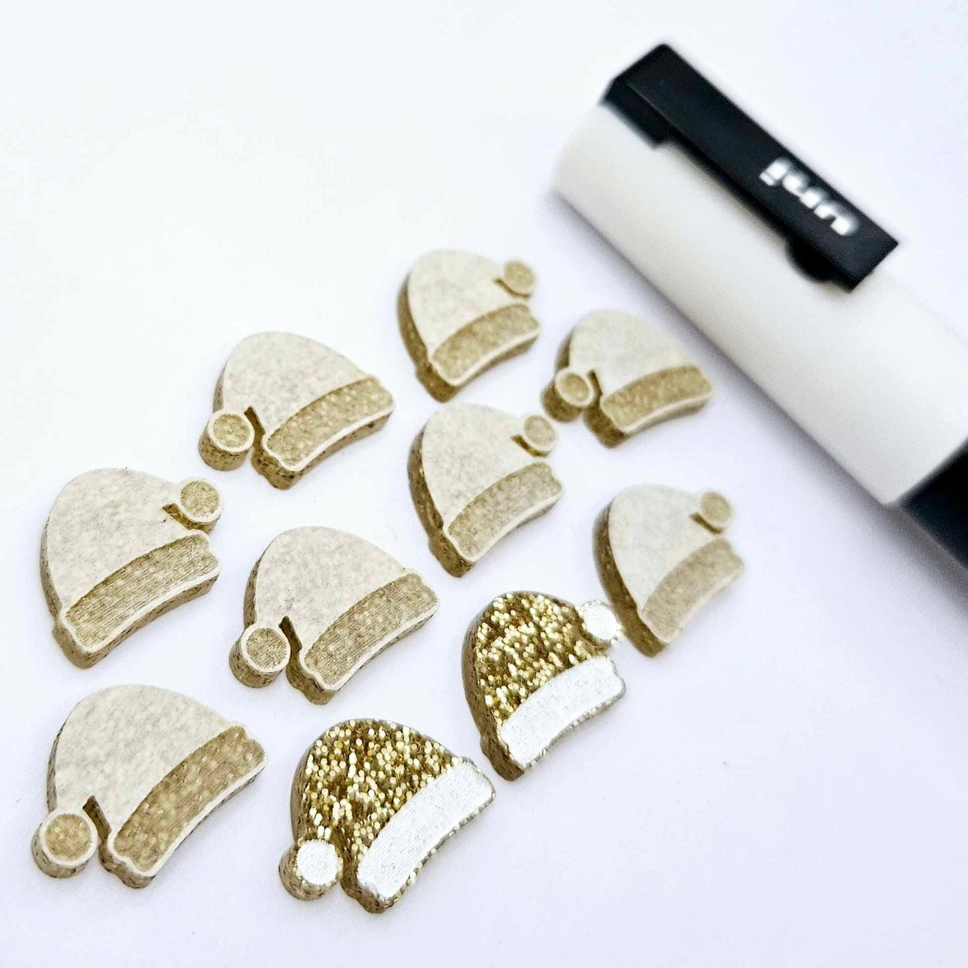 One paint-filled pair of gold glitter acrylic Santa hat stud blanks alongside multiple unpainted pieces still covered with protective paper, pictured with a White Posca Paint Pen