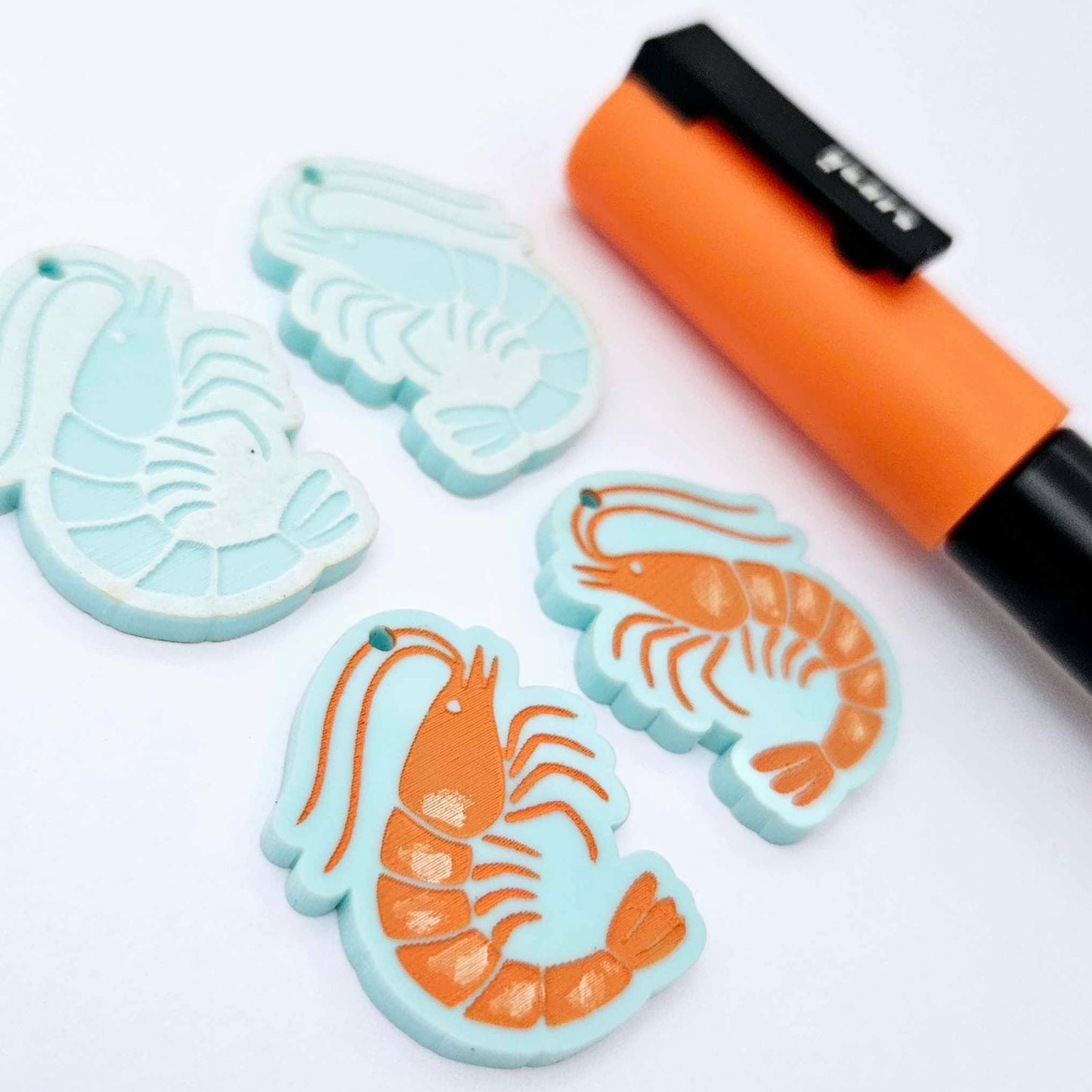 Two pairs of pastel aqua acrylic prawn dangle earring blanks with engraved details laying flat with an orange posca paint pen