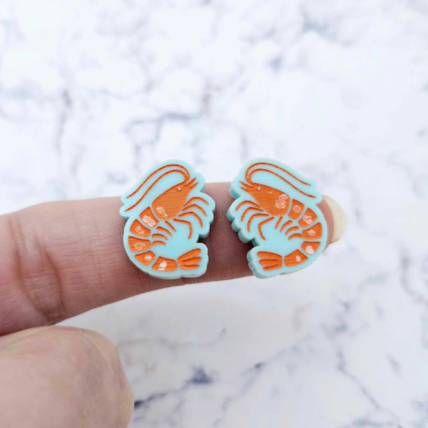 One pair of paint-filled pastel aqua acrylic prawn stud earring blanks shown on a finger for size reference