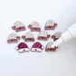 One paint-filled pair of hot pink glitter acrylic Santa hat toppers alongside multiple unpainted pieces still covered with protective paper, pictured with an uncapped White Posca Paint Pen ready for paint filling
