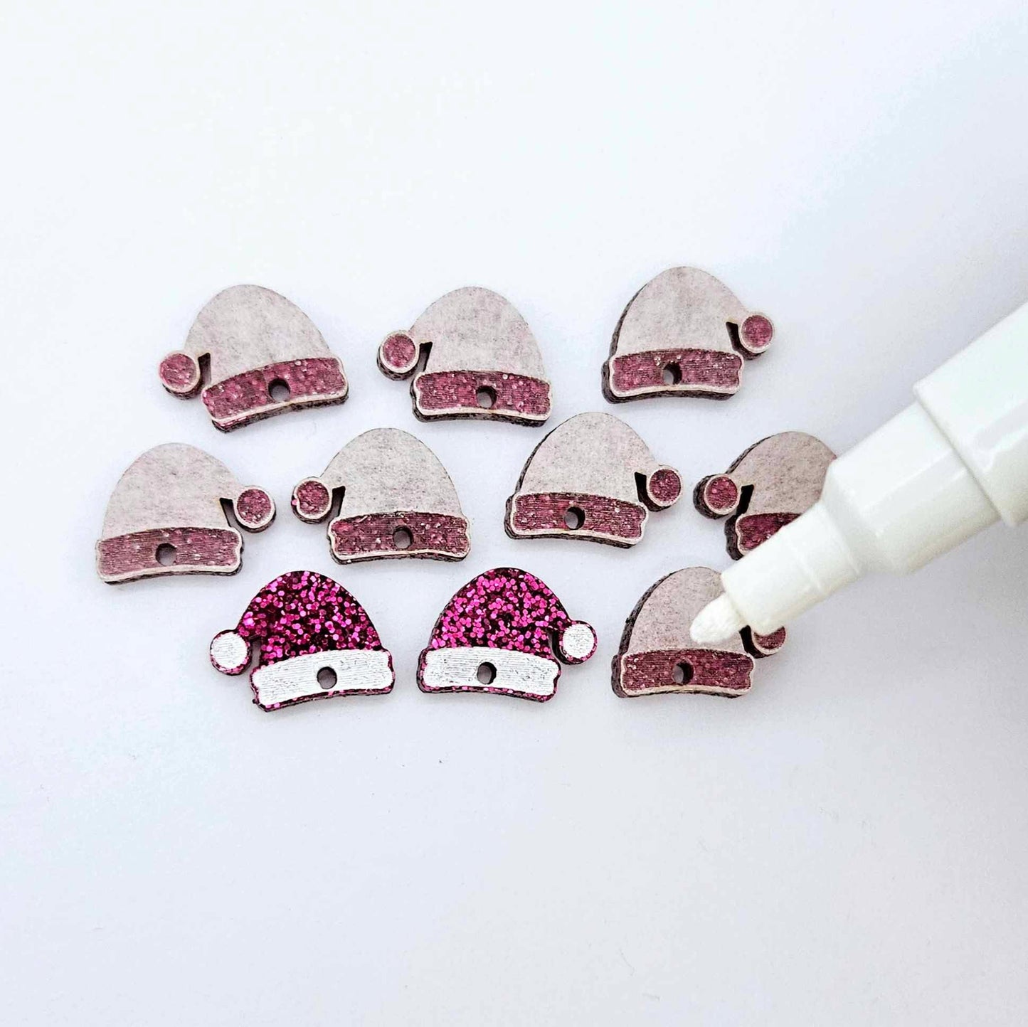 One paint-filled pair of hot pink glitter acrylic Santa hat toppers alongside multiple unpainted pieces still covered with protective paper, pictured with an uncapped White Posca Paint Pen ready for paint filling