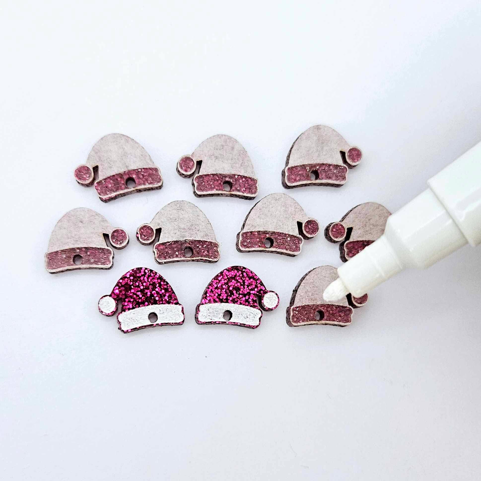 One paint-filled pair of hot pink glitter acrylic Santa hat toppers alongside multiple unpainted pieces still covered with protective paper, pictured with an uncapped White Posca Paint Pen ready for paint filling