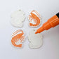 One pair of white marble prawn dangle blanks with protective paper attached, alongside one paint-filled pair with a posca paint pen uncapped ready to paint