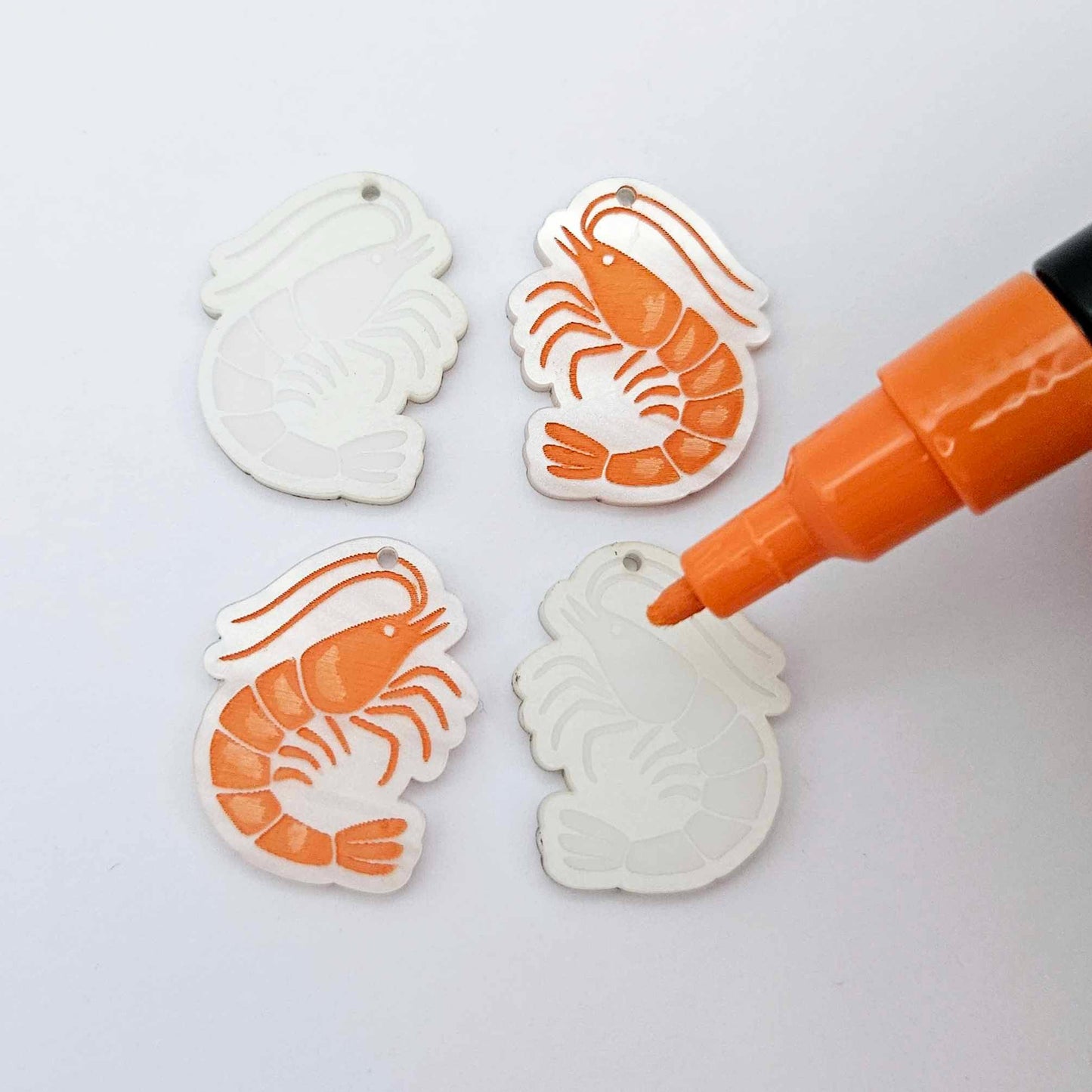 One pair of white marble prawn dangle blanks with protective paper attached, alongside one paint-filled pair with a posca paint pen uncapped ready to paint