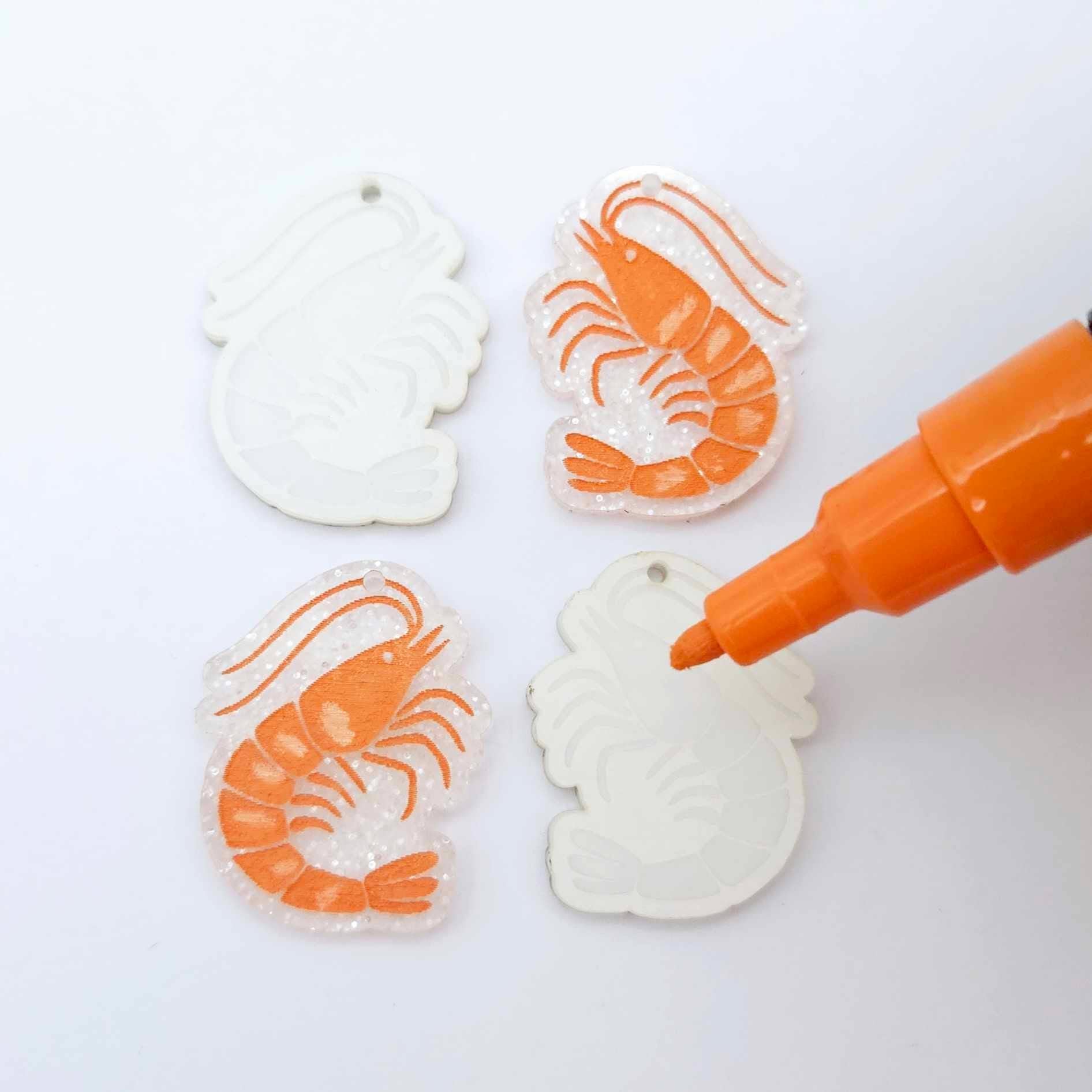 One pair of fairy dust glitter prawn dangle blanks with protective paper attached, alongside one paint-filled pair with a posca paint pen uncapped ready to paint