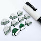 One paint-filled pair of green glitter acrylic Santa hat stud earring blanks alongside multiple unpainted pieces still covered with protective paper, pictured with a White Posca Paint Pen