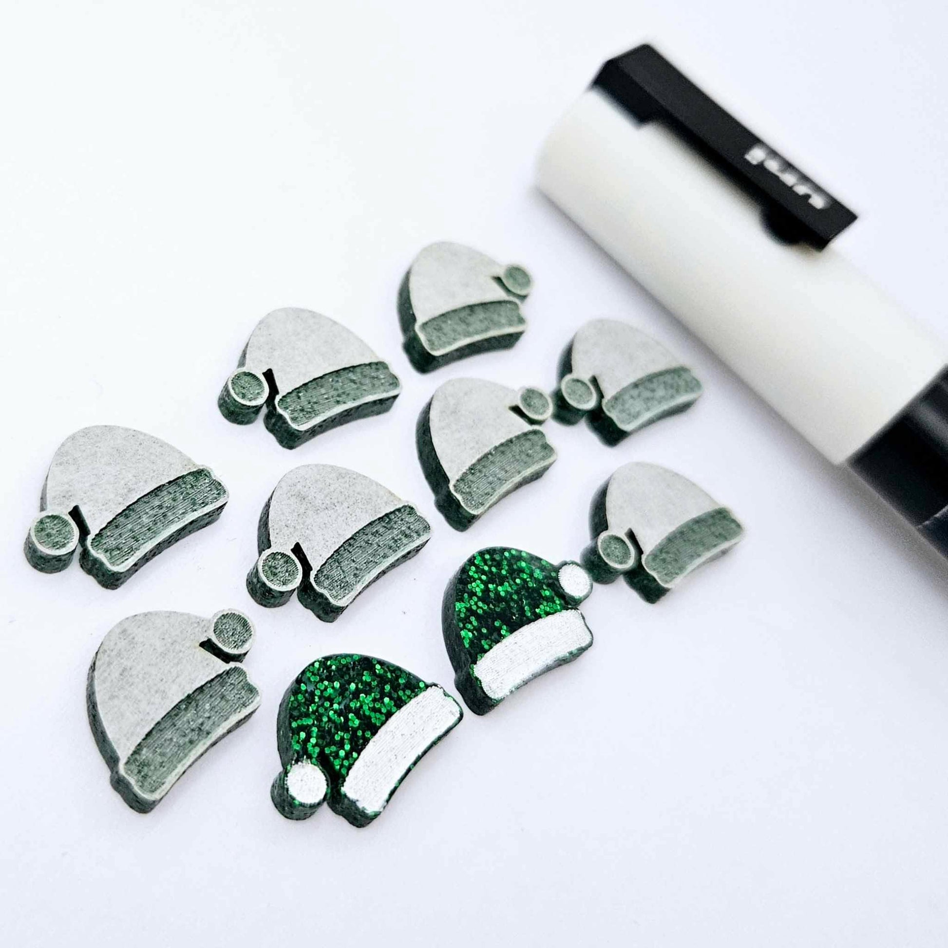 One paint-filled pair of green glitter acrylic Santa hat stud earring blanks alongside multiple unpainted pieces still covered with protective paper, pictured with a White Posca Paint Pen