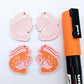 One pair of pastel pink acrylic engraved prawn dangle earring blanks with protective paper on alongside a paint filled pair, laying flat with an orange posca paint pen