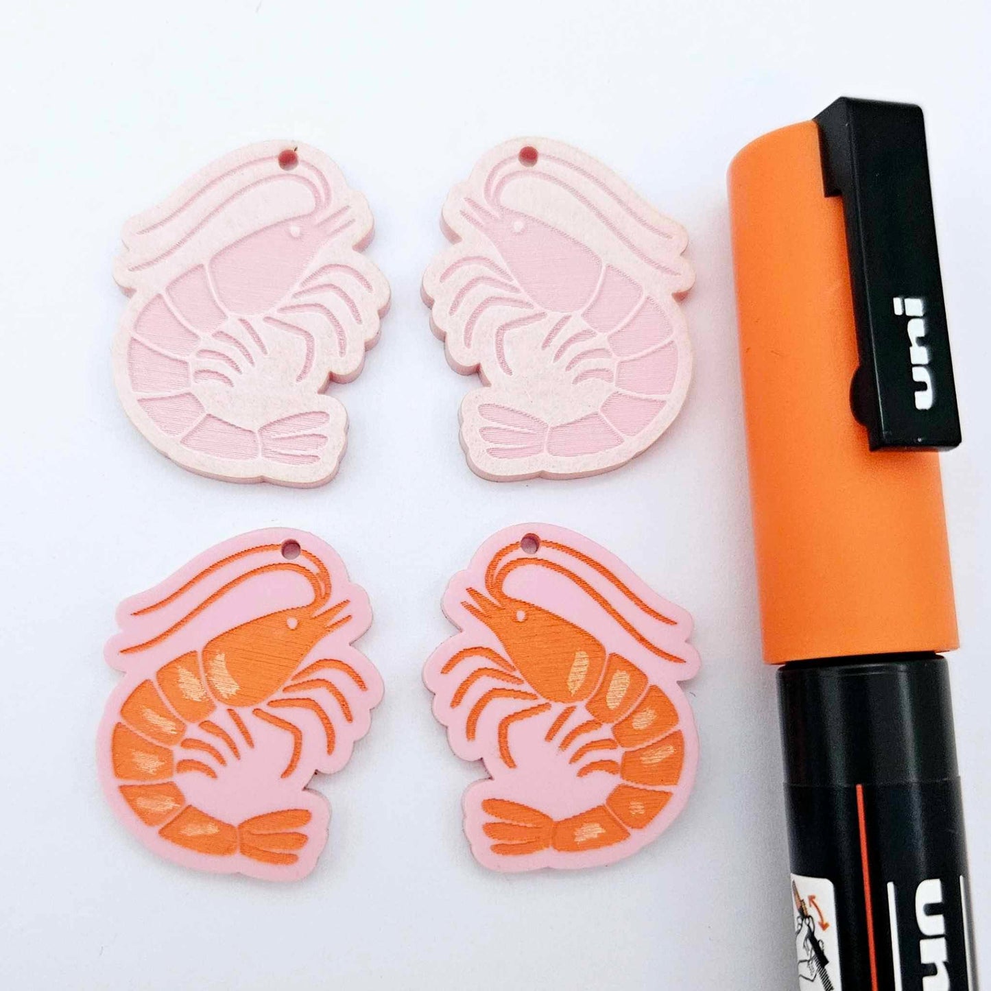 One pair of pastel pink acrylic engraved prawn dangle earring blanks with protective paper on alongside a paint filled pair, laying flat with an orange posca paint pen