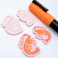One pair of pastel pink acrylic engraved prawn dangle earring blanks with protective paper on alongside a paint filled pair, laying flat with an orange posca paint pen