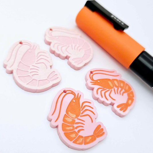 One pair of pastel pink acrylic engraved prawn dangle earring blanks with protective paper on alongside a paint filled pair, laying flat with an orange posca paint pen