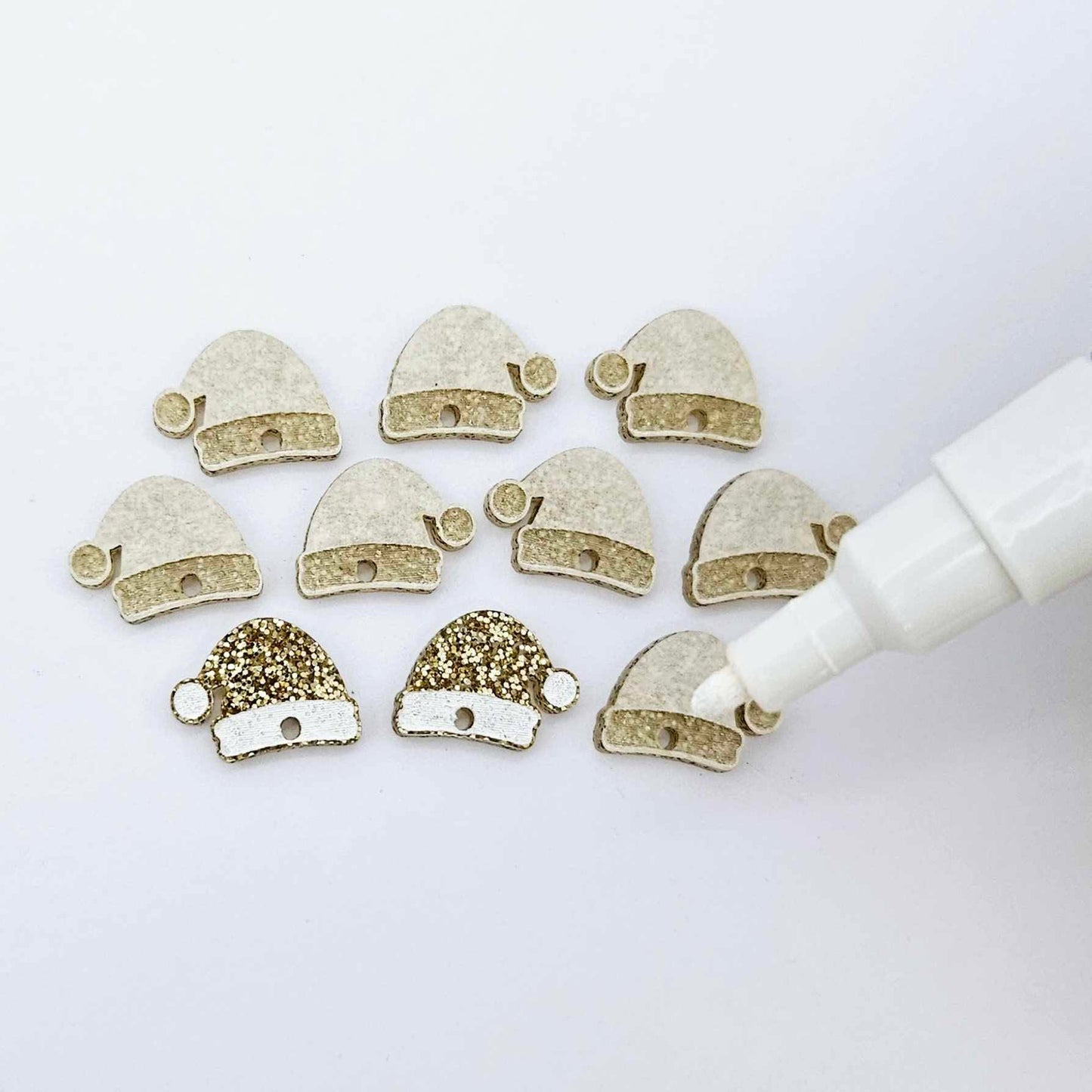 One paint-filled pair of gold glitter acrylic Santa hat toppers alongside multiple unpainted pieces still covered with protective paper, pictured with an uncapped White Posca Paint Pen ready for paint filling