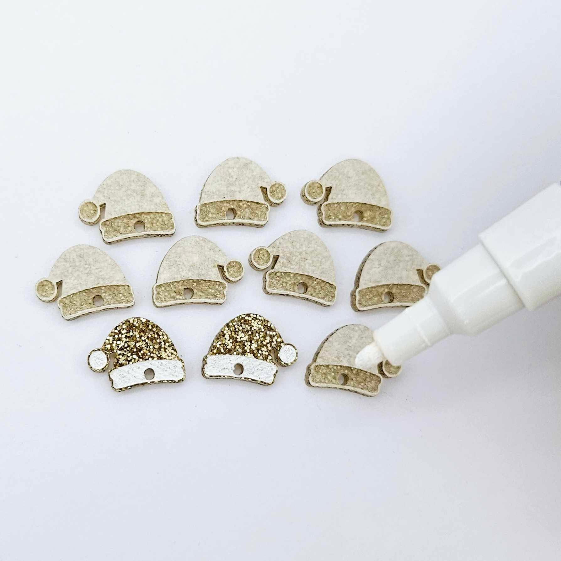One paint-filled pair of gold glitter acrylic Santa hat toppers alongside multiple unpainted pieces still covered with protective paper, pictured with an uncapped White Posca Paint Pen ready for paint filling