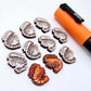 One paint-filled pair of red glitter acrylic prawn stud earring blanks with multiple unpainted pieces still covered with protective paper, pictured alongside an Orange Posca Paint Pen