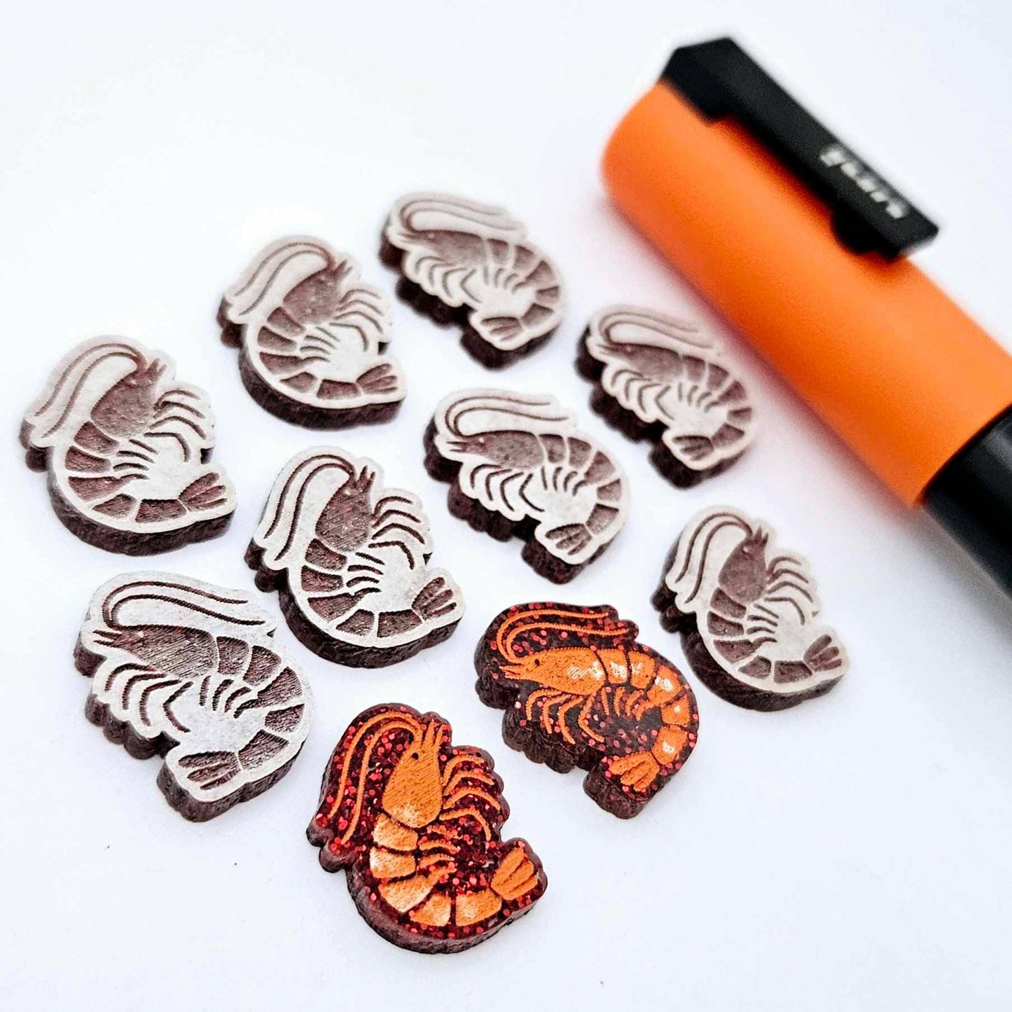 One paint-filled pair of red glitter acrylic prawn stud earring blanks with multiple unpainted pieces still covered with protective paper, pictured alongside an Orange Posca Paint Pen