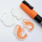 One pair of white marble acrylic engraved prawn dangle earring blanks with protective paper on alongside a paint filled pair, laying flat with an orange posca paint pen