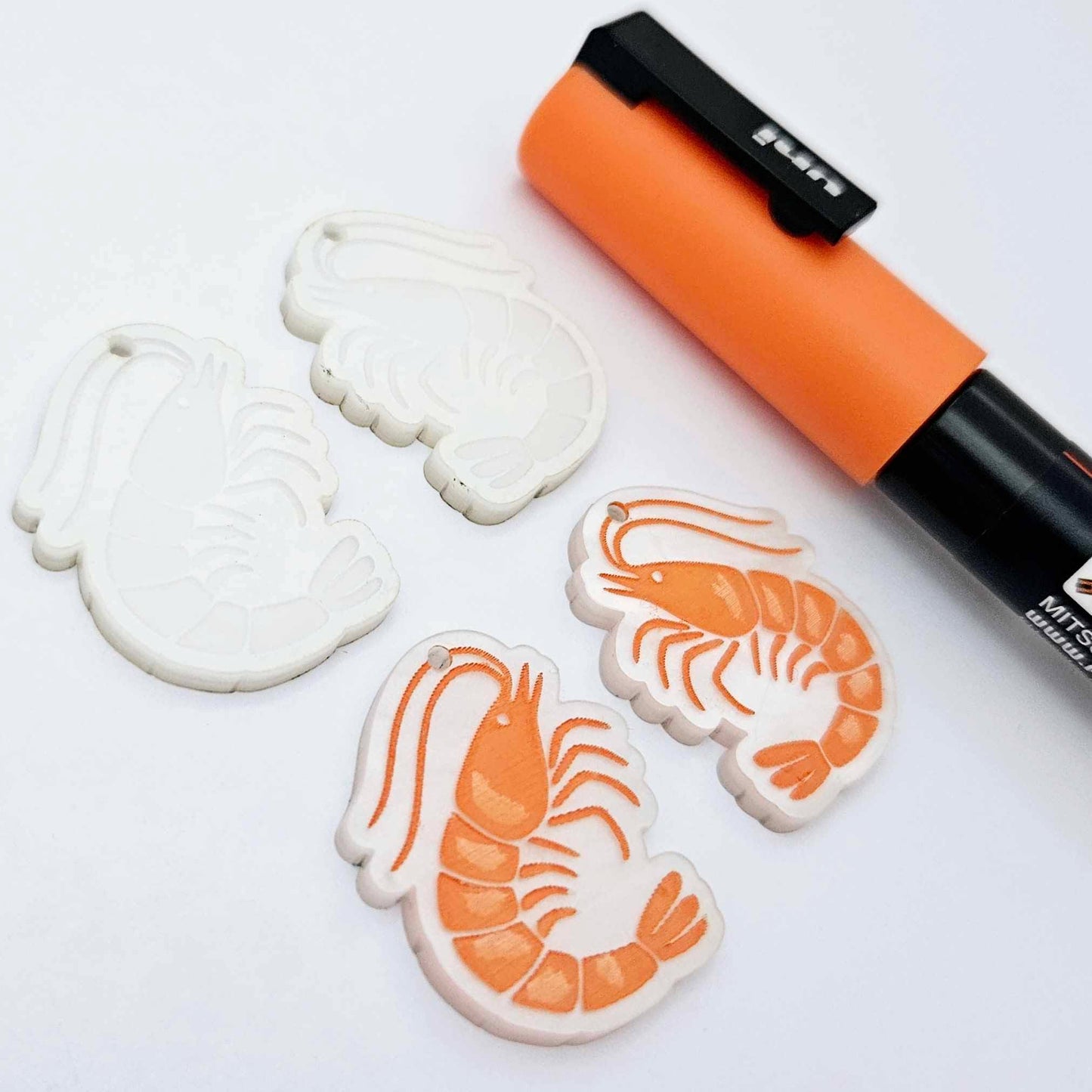 One pair of white marble acrylic engraved prawn dangle earring blanks with protective paper on alongside a paint filled pair, laying flat with an orange posca paint pen