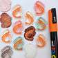 A mixture of Pastel Pink, Pastel Aqua, Red Glitter, Fairy Dust Glitter and White Marble acrylic prawn dangle earring blanks laying flat with an orange posca paint pen