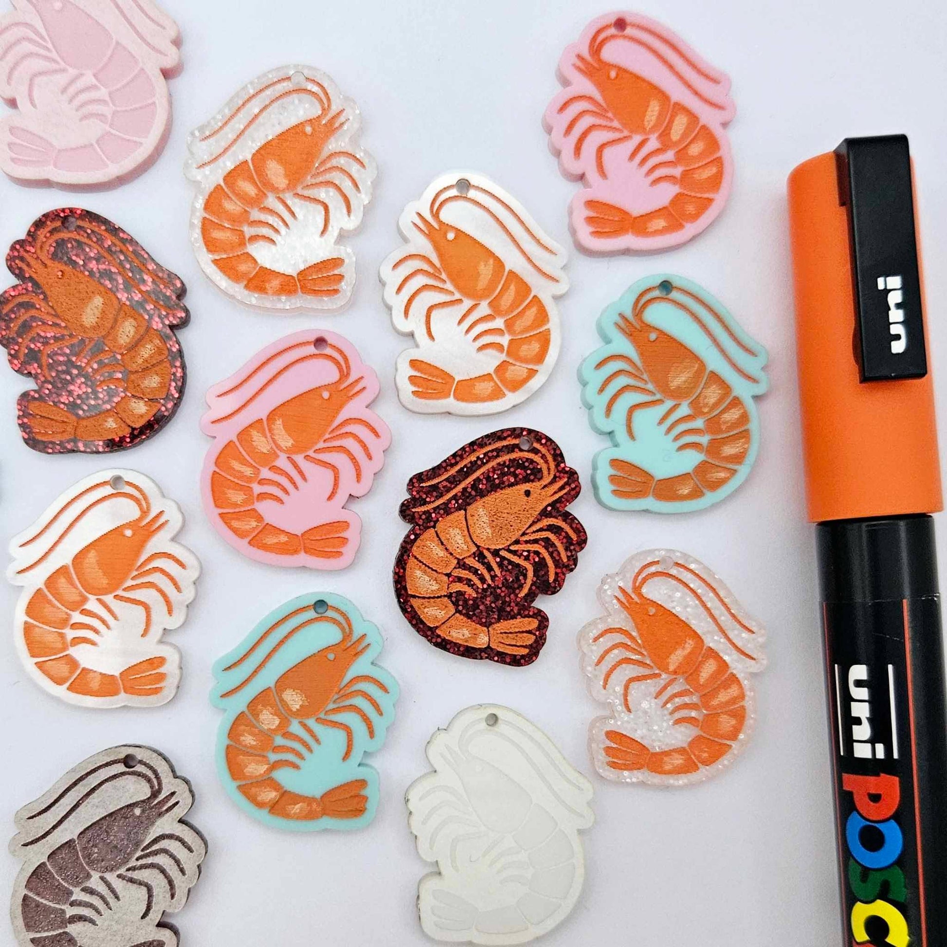 A mixture of Pastel Pink, Pastel Aqua, Red Glitter, Fairy Dust Glitter and White Marble acrylic prawn dangle earring blanks laying flat with an orange posca paint pen