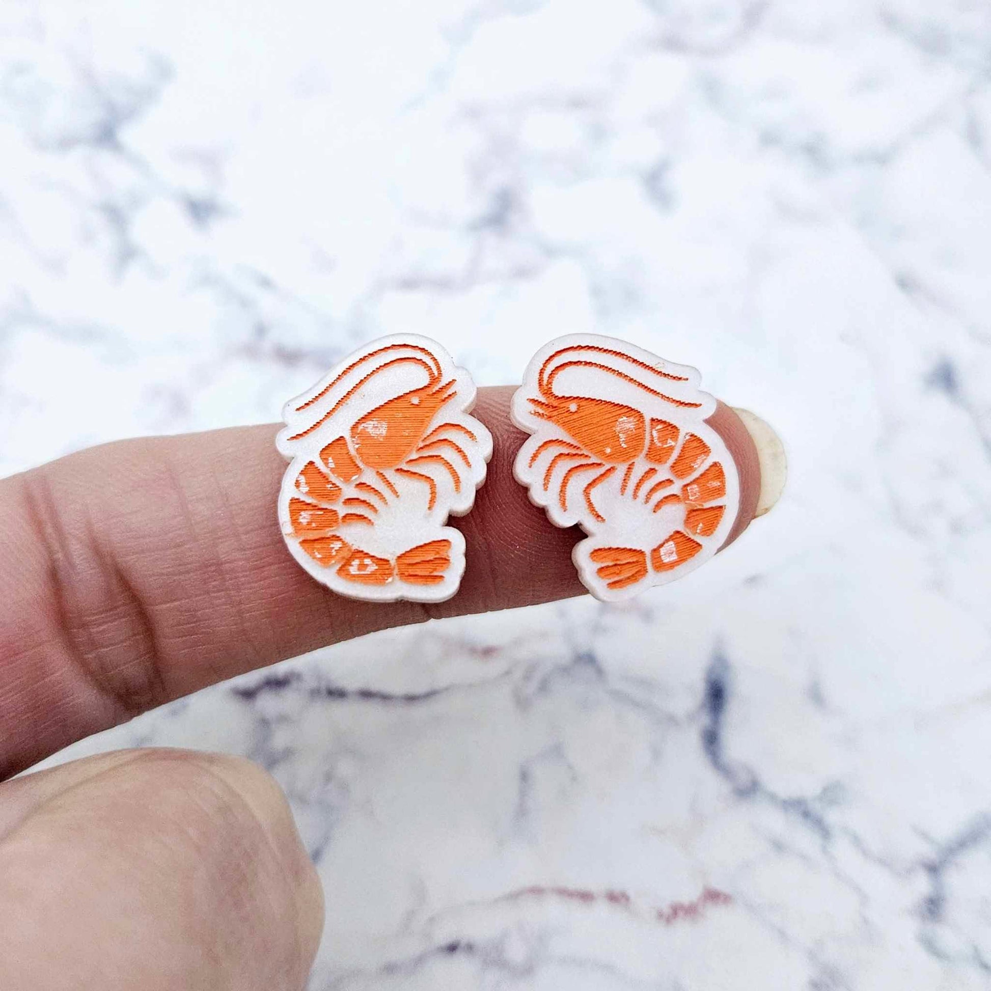 One pair of paint-filled White Marble acrylic prawn stud earring blanks shown on a finger for size reference