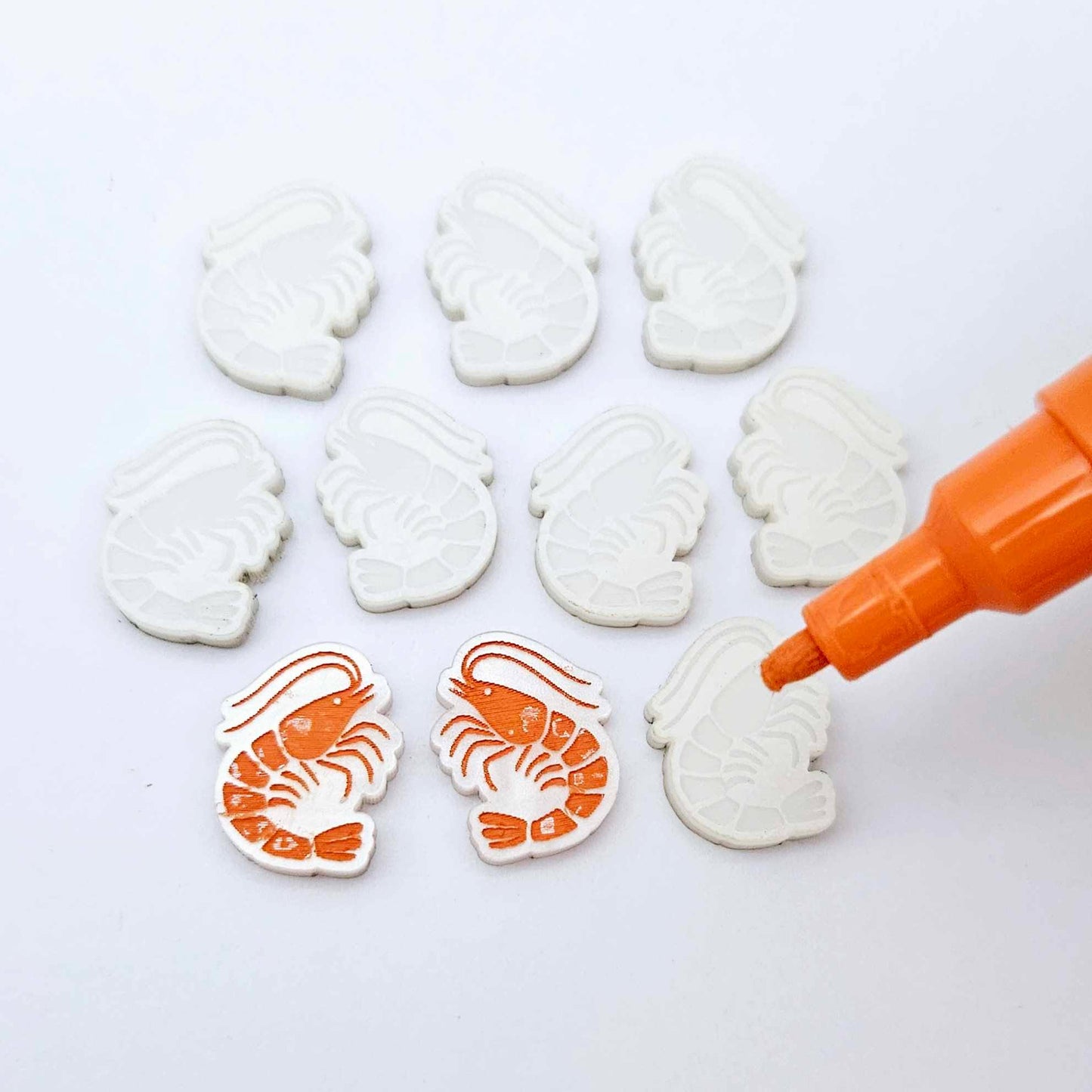 One paint-filled pair of White Marble acrylic prawn stud earring blanks with multiple unpainted pieces still covered with protective paper, pictured with an uncapped Orange Posca Paint Pen ready for paint filling