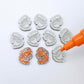 One paint-filled pair of silver glitter acrylic prawn stud earring blanks with multiple unpainted pieces still covered with protective paper, pictured with an uncapped Orange Posca Paint Pen ready for paint filling