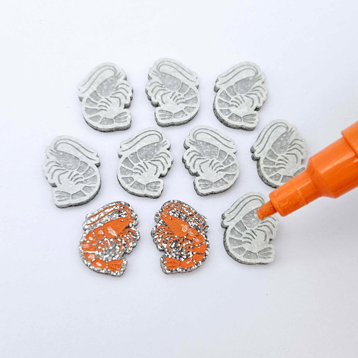 One paint-filled pair of silver glitter acrylic prawn stud earring blanks with multiple unpainted pieces still covered with protective paper, pictured with an uncapped Orange Posca Paint Pen ready for paint filling