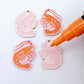 One pair of pastel pink prawn dangle blanks with protective paper attached, alongside one paint-filled pair with a posca paint pen uncapped ready to paint