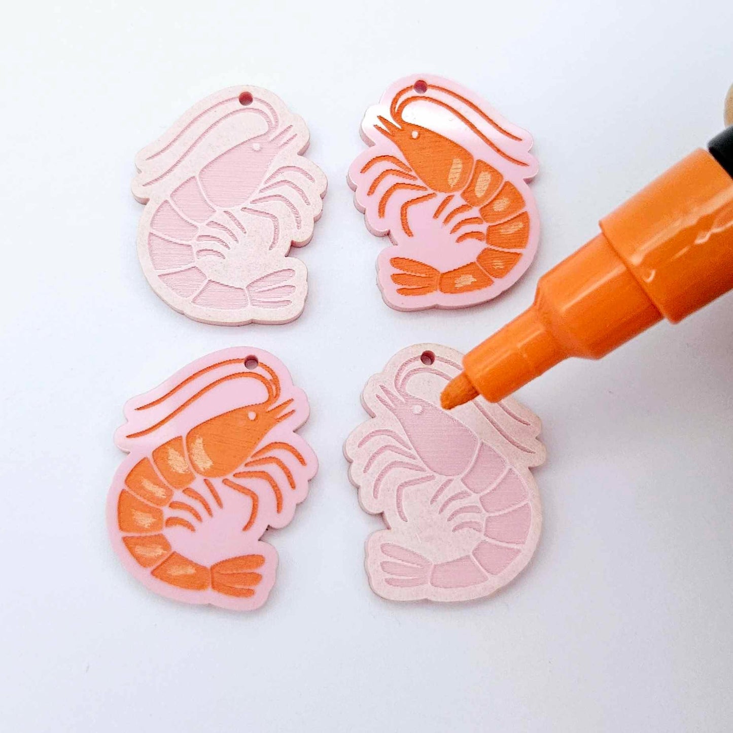 One pair of pastel pink prawn dangle blanks with protective paper attached, alongside one paint-filled pair with a posca paint pen uncapped ready to paint