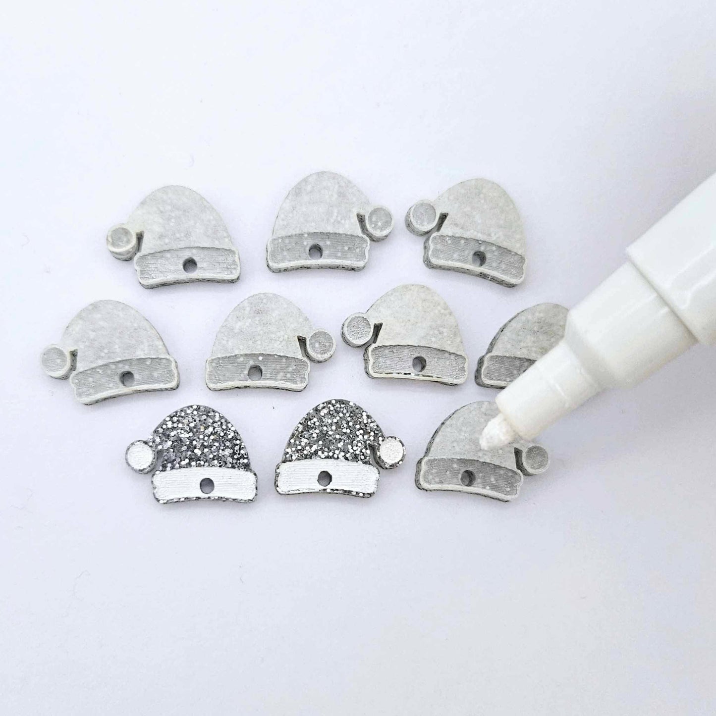 One paint-filled pair of silver glitter acrylic Santa hat toppers alongside multiple unpainted pieces still covered with protective paper, pictured with an uncapped White Posca Paint Pen ready for paint filling