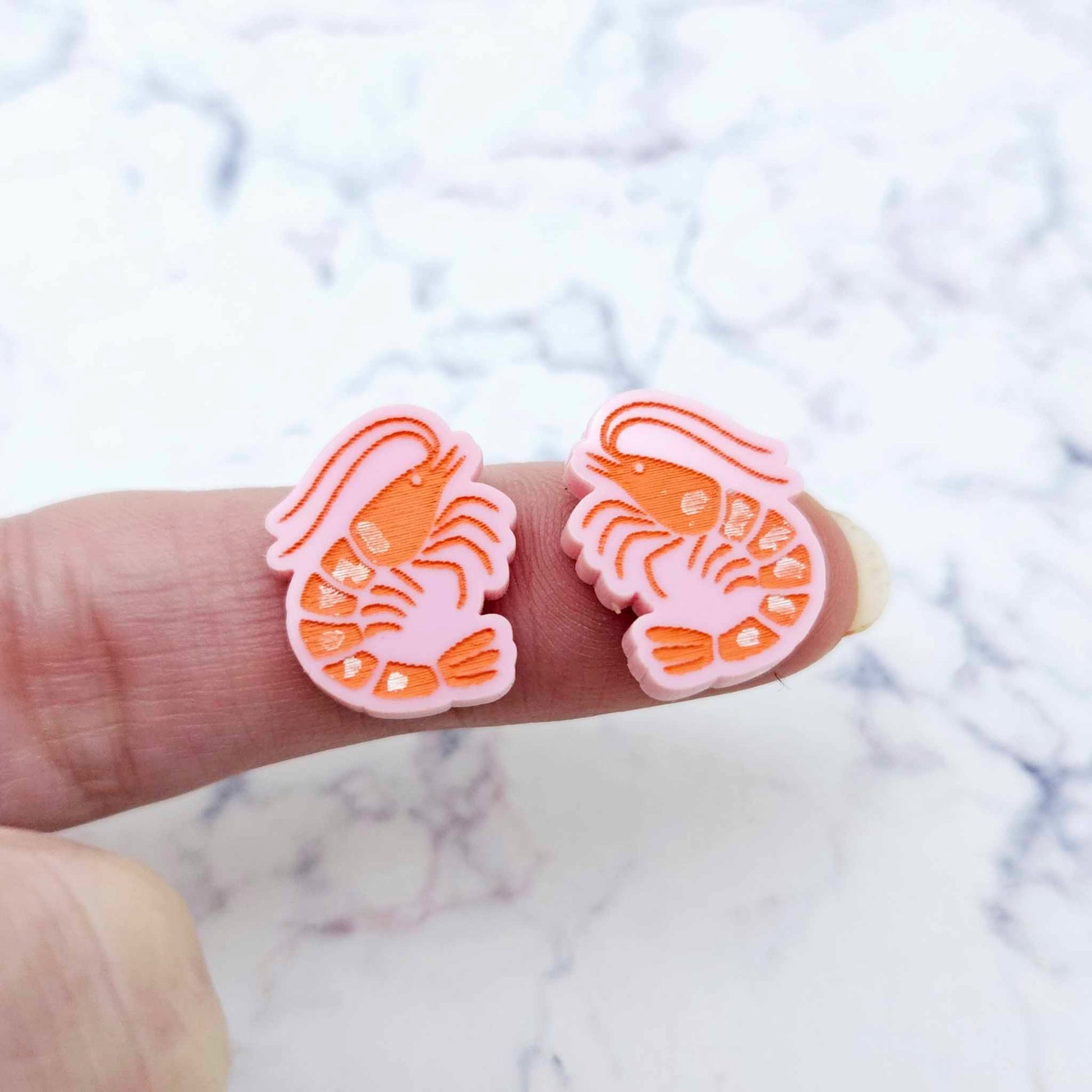One pair of paint-filled pastel pink acrylic prawn stud earring blanks shown on a finger for size reference