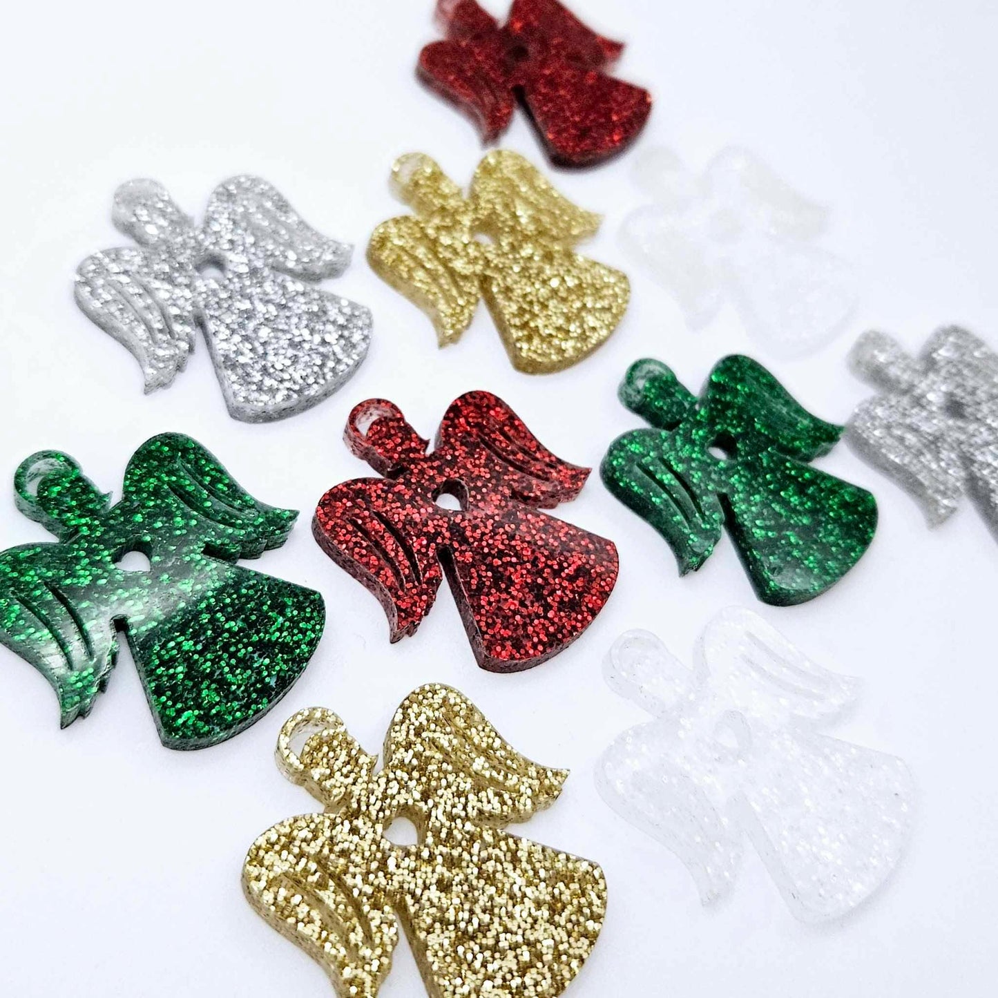 A mixture of gold glitter, red glitter, green glitter, silver glitter and Fairy Dust Glitter acrylic 32mm angel dangle earring blanks laying flat on a white background